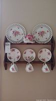 Front view of two-tier wooden shelf with china plates, saucers, teacups, and a teapot arranged and hanging on hooks.