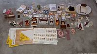 Wide view of dollhouse miniatures and furniture pieces, assorted packages of miniature furniture sets, plastic figures, and flooring sheets laid out on a surface.