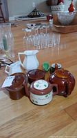 Photo showing brown and white pottery items including Brown Betty teapot, Irish teapot, ironstone pitchers, and pottery with lids. Also shows clear glassware in background.