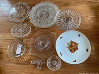 Top view of 11 glass serving pieces and 1 China cake plate with fruit design arranged on floor wooden surface