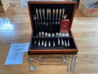 Wooden box containing Gorham Silverplate Invitation pattern flatware with multiple utensils including forks, knives, spoons, and additional decorative spoons and forks laid in front on wooden floor.