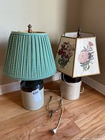 Photo showing both lamps and the extra lampshade frame on wooden floor against a beige wall. The lamps have ceramic jug bases with different lampshades.