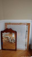 Two mirrors leaning against a wall, one smaller with a dark reddish-brown scalloped wood frame in front, a larger natural wood framed mirror partially wrapped in bubble wrap behind it.