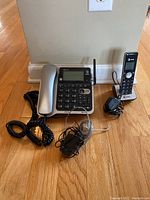 Corded and cordless AT&T telephones with cords and power adapters placed on hardwood floor against wall.