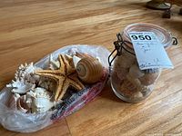 Photo of assorted sea shells and starfish next to a glass jar filled with small shells on a wooden floor.