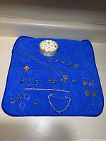 Jewelry displayed on blue cloth including rings, bracelets, earrings, cross pendant, necklace, and decorative small round tin box.