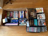 Box containing multiple VHS tapes, DVDs, CDs, cassettes, and two books, all assorted and loosely packed.