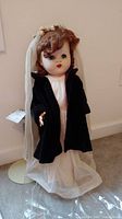 Full view of vintage Fairyland doll in black velvet coat, white dotted flocked gown, veil, and stand with plastic cover on carpeted floor