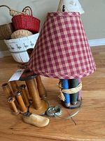 Photo showing the lamp with a base constructed of large wooden spools wrapped with twine and a red and beige checkered fabric lampshade. Nearby are several vintage wooden spools and a wooden shoe form.