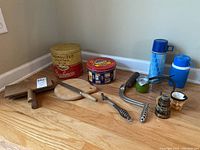 Overview of all items in the lot arranged on wood floor, shows tins, thermos, wooden hook, chimney cranks, glass and hooks