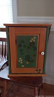 Front angled view of orange and green painted wooden wall cabinet with floral design on door, placed on chair in room with window background.