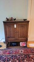 Full front view of wood cabinet showing two carved doors, top edge trim, small items on top and some books and electronic device below. Cabinet stands on four legs with front footrest.