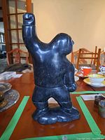 Front view of dark grey-green soapstone carving of Inuit figure raising one arm, standing on base.