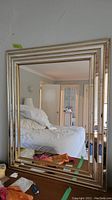 Full front view of the large rectangular mirror reflecting a bedroom. Mirror has a gold toned multi-layered frame with alternating mirrored and decorative gold block sections.