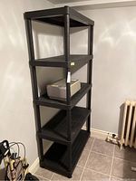 Full view of the black plastic shelving unit standing against a wall with a small device on one shelf, showing overall size and setup.