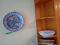 Ceramic wall plate mounted on wall showing blue fish design with leaf border and white bowl on shelf below.