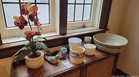 Set of artisan pottery, planters, bird figurines, and faux floral arrangement displayed on wooden cabinet near window.