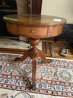 Full view of the round wooden side table showing its shape, drawer and pedestal base with claw foot metal casters.