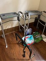Two aluminum Guardian Signature walkers with attached gray plastic tray tables, gray padded handles, standing on wooden floor with incontinence product packages nearby.