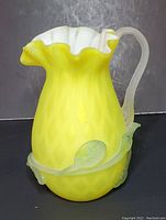 Front angled view of the yellow satin Mt Washington glass pitcher, showing ruffled white interior rim, frosted handle, and applied leaves.