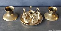 Front-angle view of brass swan taper candle holder and two matching round brass candle holders on flat surface.