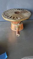 Brass and copper sterno warmer grill with slotted circular brass top and wooden handle, shown from above and front perspective.