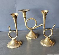 Photo showing three brass equestrian hunting horn taper candle holders on a dark surface against a gray backdrop, positioned in a triangle formation highlighting their horn shape and base details.