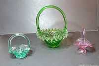 Three glass baskets on gray surface and backdrop, showing size and colors of the lot including the tall green hobnail basket and two smaller unmarked ones.