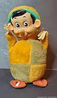 Front view of Gunderful Gund Pinocchio plush baby toy showing yellow and green patchwork body, molded head with facial details, and yellow hat.