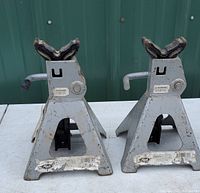 Pair of gray Mack 3 ton floor jack stands with visible wear and rust, set on white surface with a green vertical panel background.