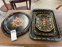 Five toleware trays arranged on table showing size and shape variety, three rectangular stacked and two circular placed side by side. Hand-painted floral decoration on black background. Visible scratches.