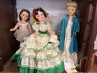 Lot showing three vintage dolls standing next to each other on a shelf: ballerina doll, Madame Alexander doll in detailed floral dress and bonnet, doll in white dress and blue coat.