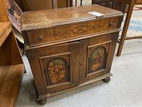 Front of antique wooden cabinet showing drawer with floral knobs and hand-painted Gypsy-themed portraits on each door, with visible wear and scratches on wood.
