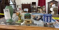 View of entire lot including ceramic vase, bird figure, metal urn, cloisonné jar, blue patterned bowl, and tea cup and saucer on glass table