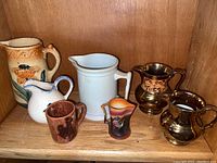 Six pitchers in different sizes arranged on a wooden shelf; includes copper luster pitchers, white ceramic pitcher, larger earth tone ceramic pitcher, small white and blue pitcher, and small cowboy-themed ceramic mug.
