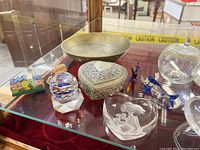 Photo showing a selection of glass paperweights, a metal trinket box, a metal bowl, and small colorful glass figurines on a glass shelf.