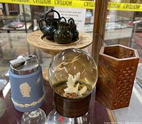 Group photo of all lot items on glass surface including lighter, paperweight, pencil holder, miniature tea set on wooden pedestal tray