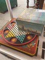 Top view photo of one Chinese Checkers game board with glass marbles on the board surface and a decorative metal box with London landmarks design.