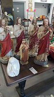 Photo showing four statues of Jesus Christ with Sacred Heart and smaller religious statue of a saint holding a child, along with a white vase or bust on a dark wooden table.