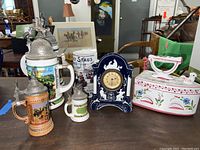 Lot shows five hand-painted ceramic beer steins with pewter lids grouped around a dark blue ceramic clock in ceramic case. Steins have varied colors and detailed scenes, clock has white decorative accents and central clock face.