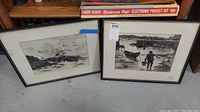 Two framed monochromatic artworks with sea scenes placed standing on the ground with shelf behind. One shows rough sea with rocky coast and clouds, other shows shoreline with boats and fisherman.