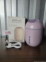 Pink bottle-shaped humidifier with USB charging cable and original box placed on a flat surface against a metal paneled background. Includes label with lot number.