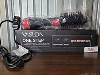 Vaslon One Step hair dryer and styler hot air brush shown in original box with power cord and control switch visible. Device is wrapped in protective plastic.