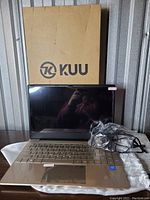 Gold KUU laptop with charger in front of KUU cardboard box, keyboard and screen visible.