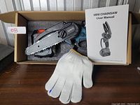 Photo of a mini chainsaw in a box next to a user manual and a pair of white gloves.