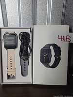 Smart watch inside original box with black and beige wristbands and charger cable visible