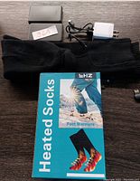 Photo showing the black heated socks, black rectangular rechargeable battery pack, USB charging cable, and white power adapter on a wooden surface along with the packaging box laid flat.
