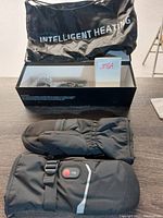 Full view of pair of black heated mitts with storage bag labeled 'Intelligent Heating' and box, showing mitts and accessories.