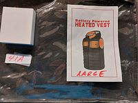 Photo showing the packaged black heated vest, labeled 'Battery Powered HEATED VEST LARGE' with a manual illustration of the vest.
