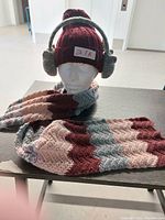 Photo shows a mannequin head displaying a burgundy knit hat with attached gray fuzzy earmuffs and a matching multicolor chevron knit scarf folded in front.